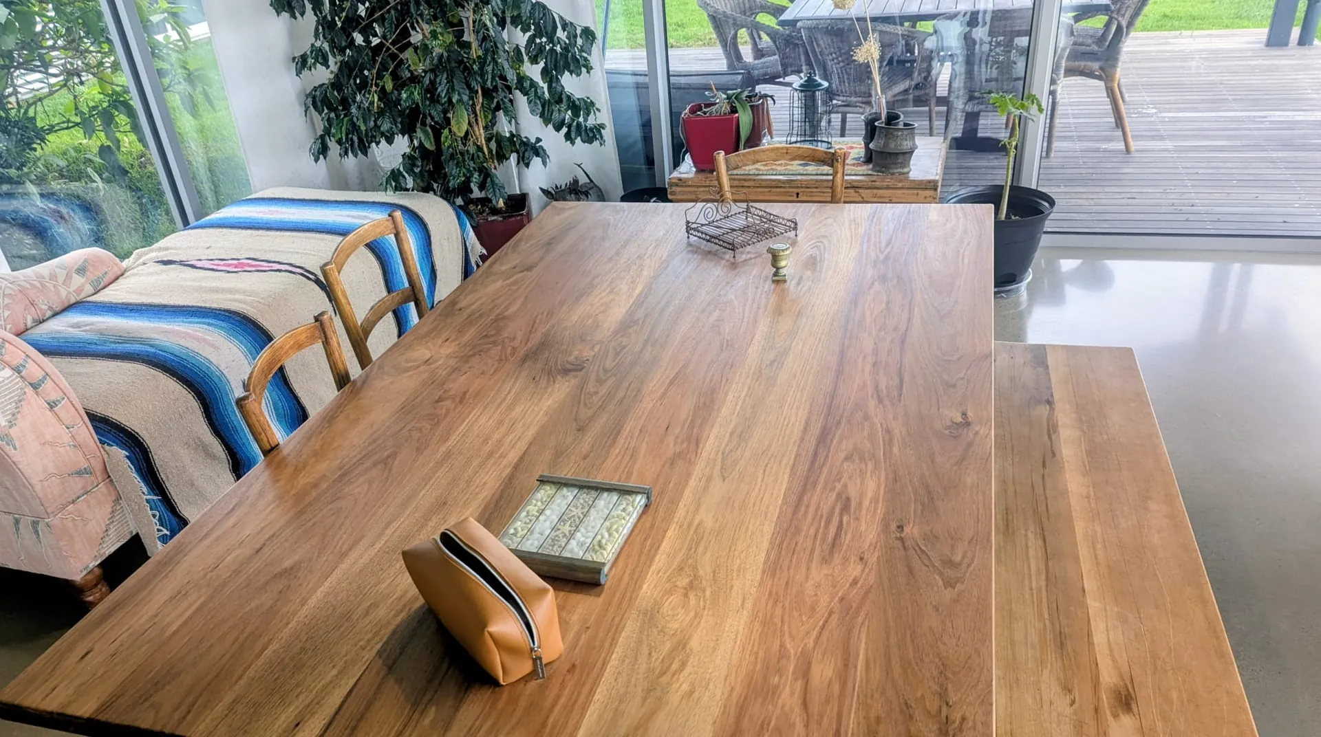 A large solid timber dining table with a matching bench in a light-filled room, looking out to a garden